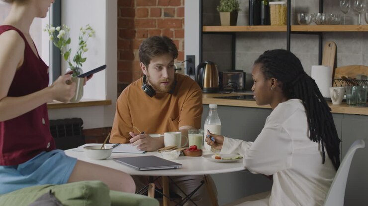 Three Roommates Having Breakfast, Using Smartphone And Taking Notes In The Kitchen