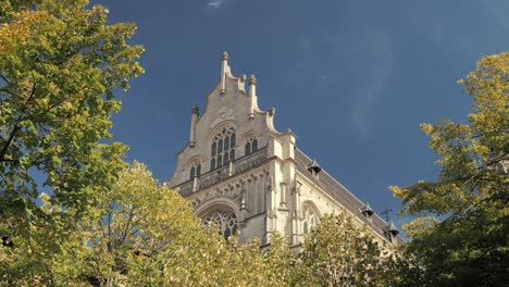 A grand Gothic-style building emerges from behind lush trees under a clear blue sky on a sunny day