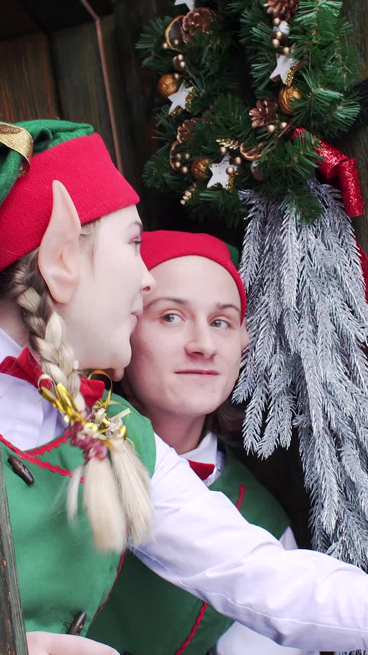 2 elves in green hats peep out of the door of the Santa Claus residence's wooden house in surprise. Vertical video