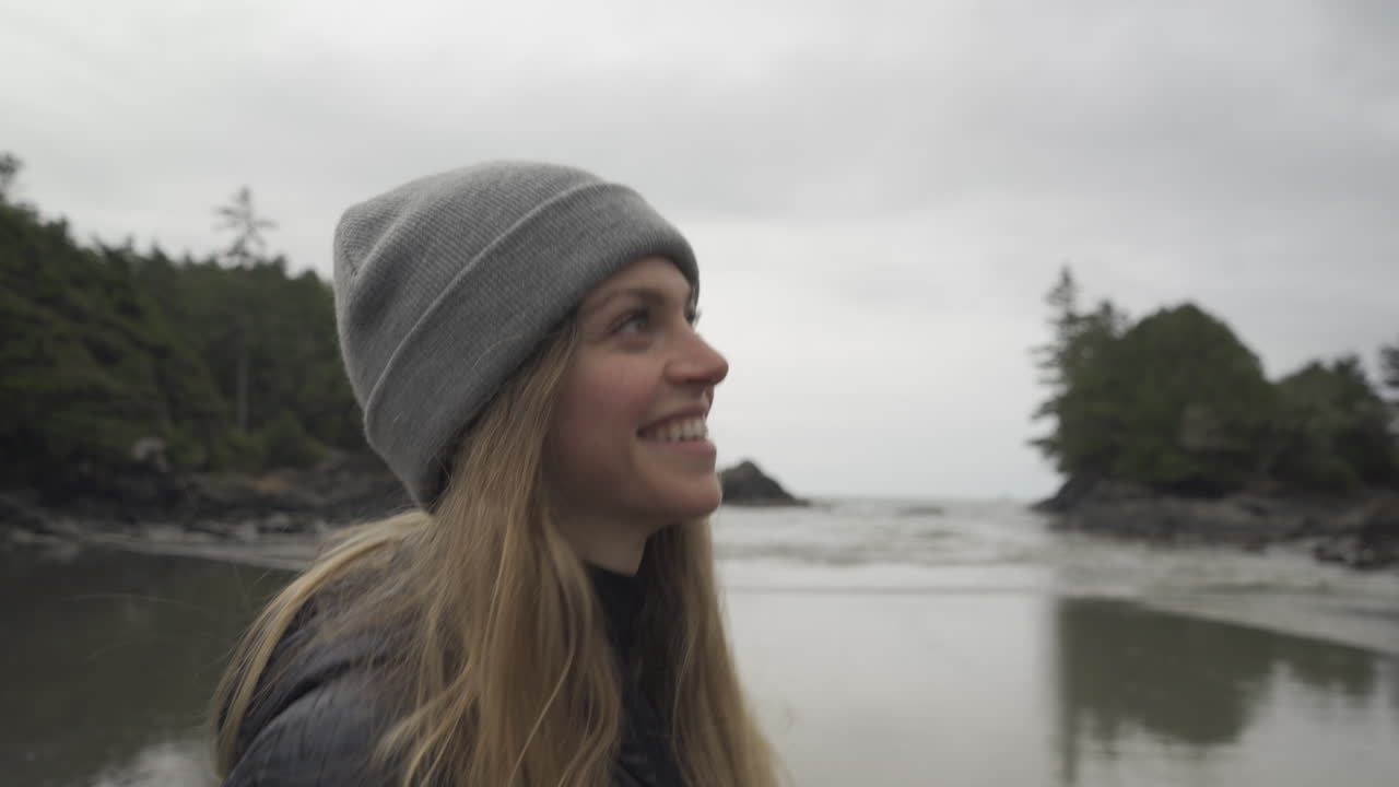 un primer plano de una chica sonriendo y caminando por la playa en tofino, columbia británica, canadá, en una mañana nublada con olas entrando en la arena.