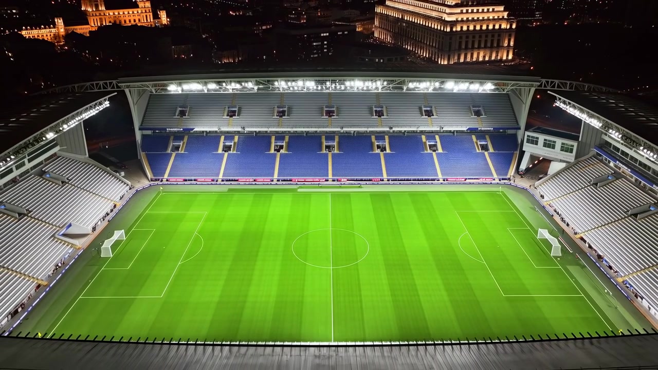 Ampia veduta di un campo da calcio vuoto all'interno di uno stadio illuminato, con file di sedili e strutture circostanti visibili di notte, che evidenzia il manto erboso verde, una composizione equilibrata e l'ambiente sereno dello stadio.