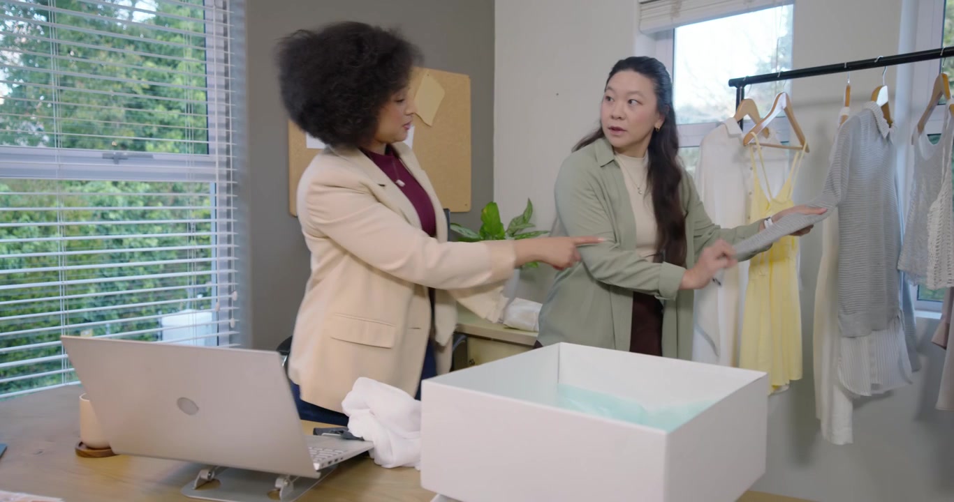 African American and Asian women unpacking box, checking laptop, comparing swatches at studio desk