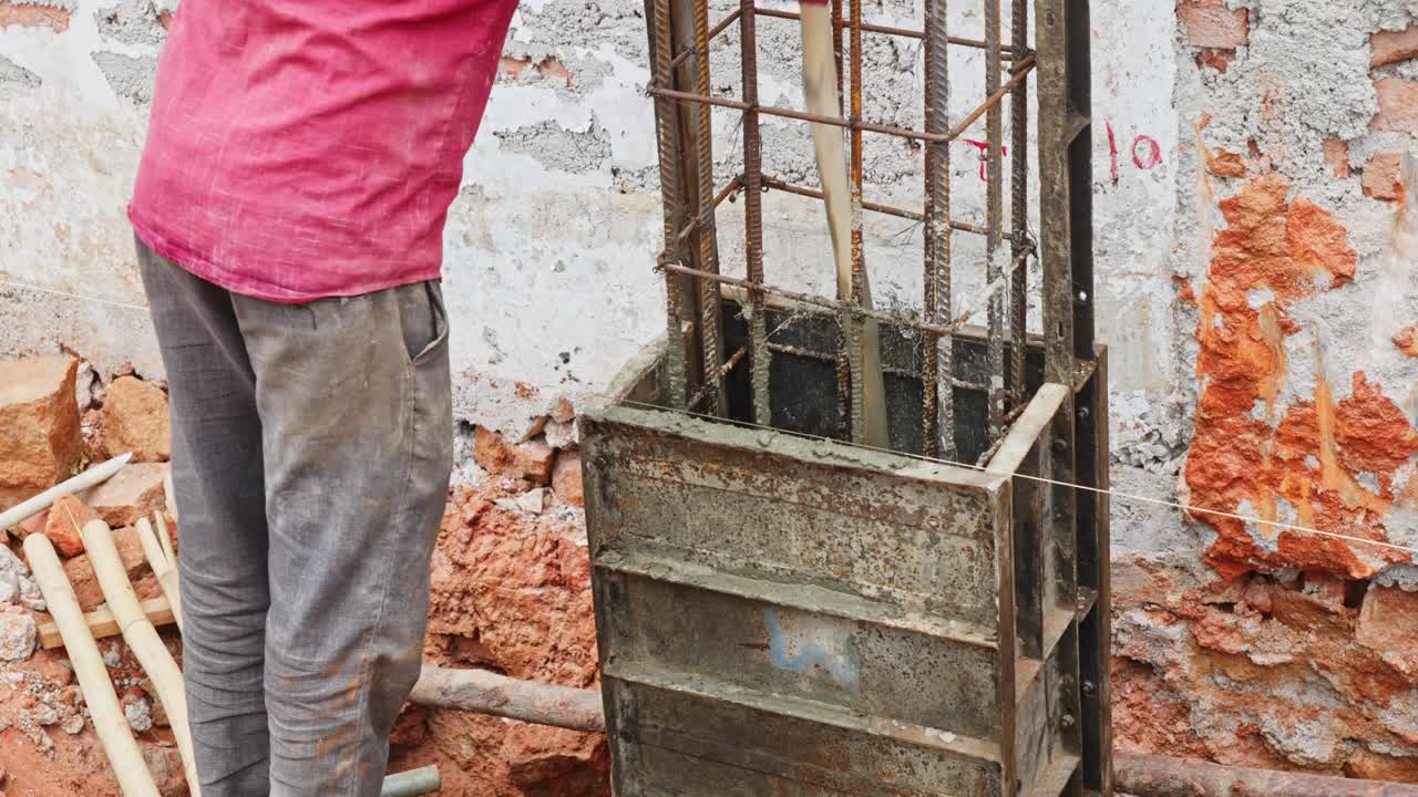 Un albañil vertiendo mezcla de hormigón en una caja de encofrado para crear una columna en una obra. Toma estable a 4K durante el día.
