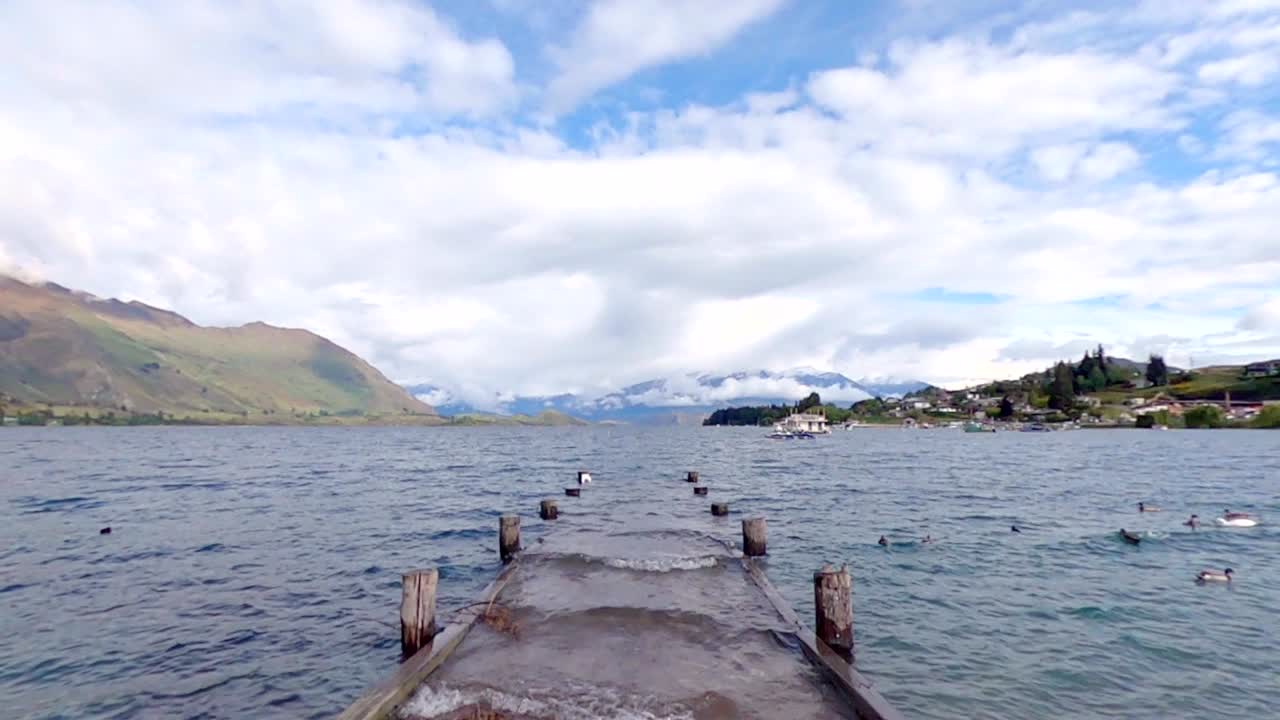 vista pacífica del lago wanaka con muelle sumergido, patos, botes y montañas en la escena