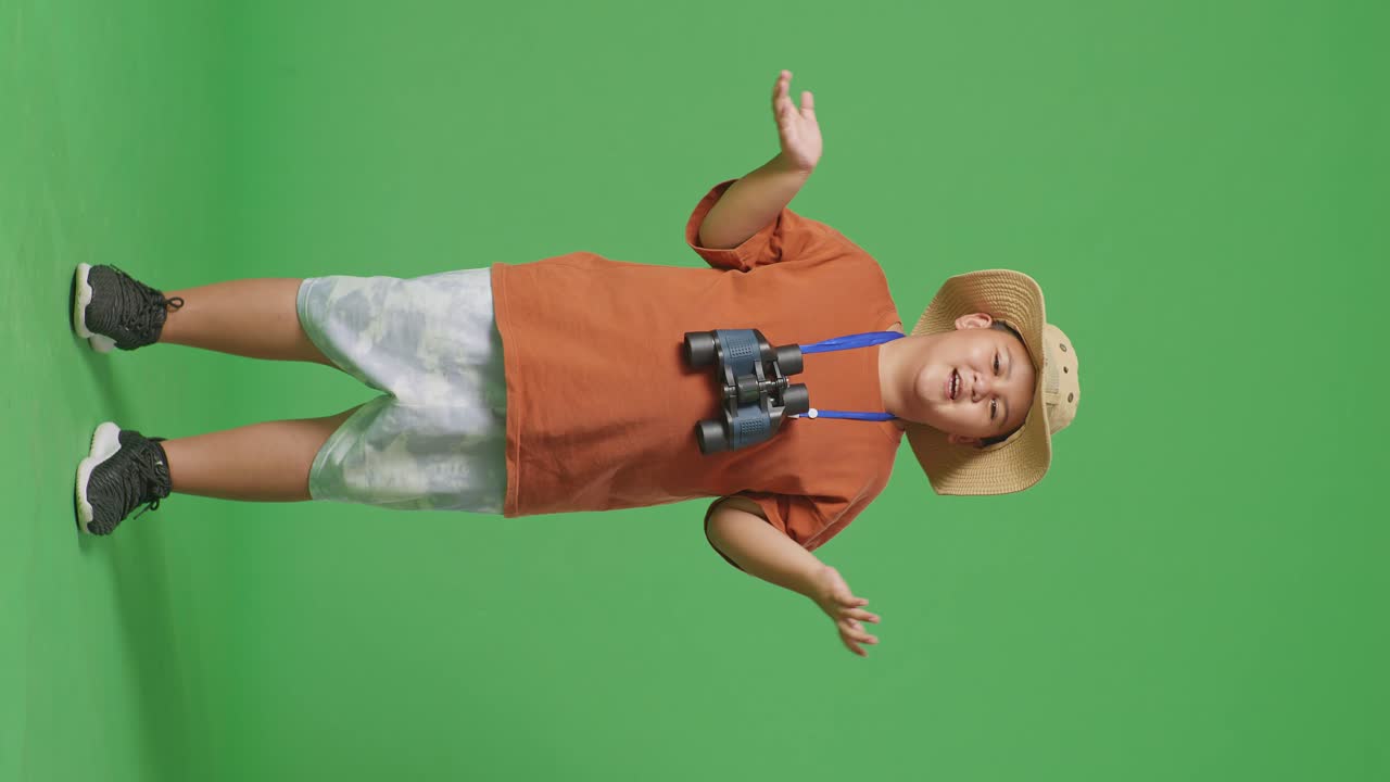 Asian Tourist Boy With A Hat Standing Doubtfully And Asking Why After Looking Through The Binoculars. Boy Researcher Examines Something On The Green Screen Background, Travel Adventure, Full Body
