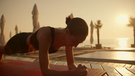 Una-Chica-Morena-Con-Cabello-Estrellado-Y-Un-Traje-Deportivo-Negro-De-Verano-Hace-Ejercicios-De-Plancha-Sobre-Una-Alfombra-Roja-En-Una-Playa-Soleada-Al-Amanecer.-Playa-Soleada-Cerrada-Con-Tablas