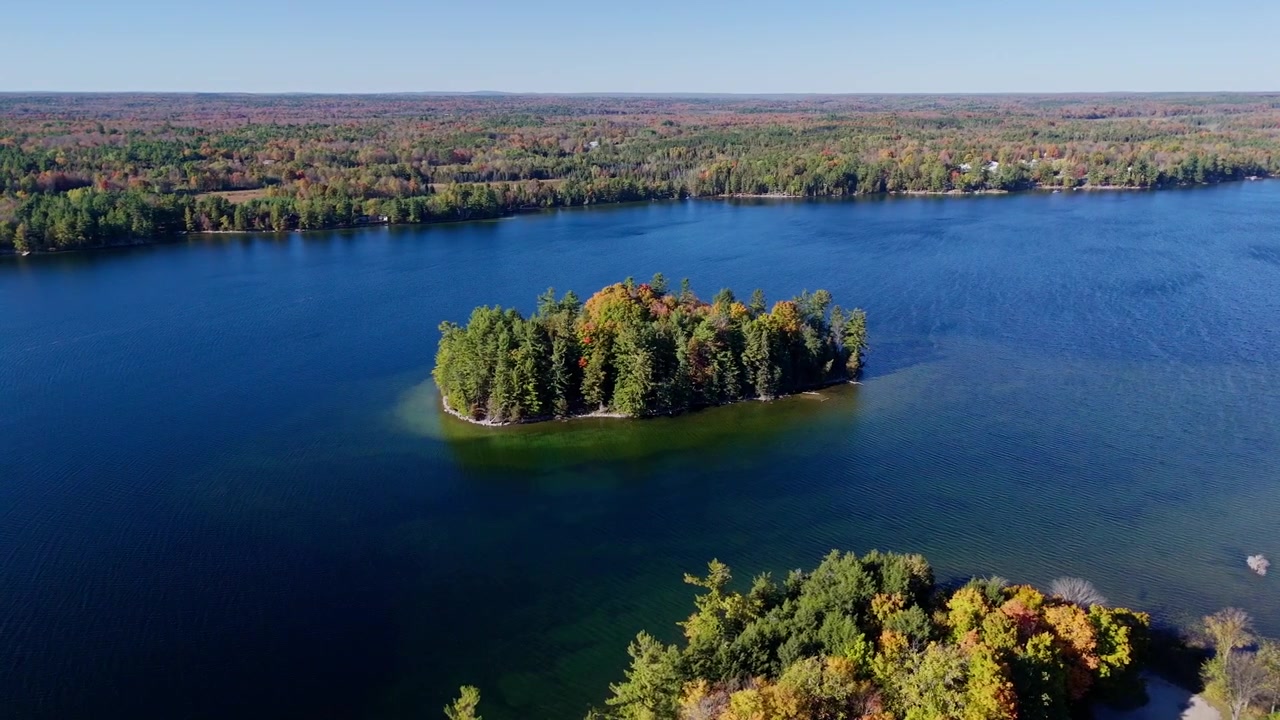 Tomada cinematográfica de drone voando lateralmente por uma pequena ilha isolada com folhagem colorida de outono em Ontário. Ondulações visíveis do vento no lago e belas cores de outono na natureza selvagem.
