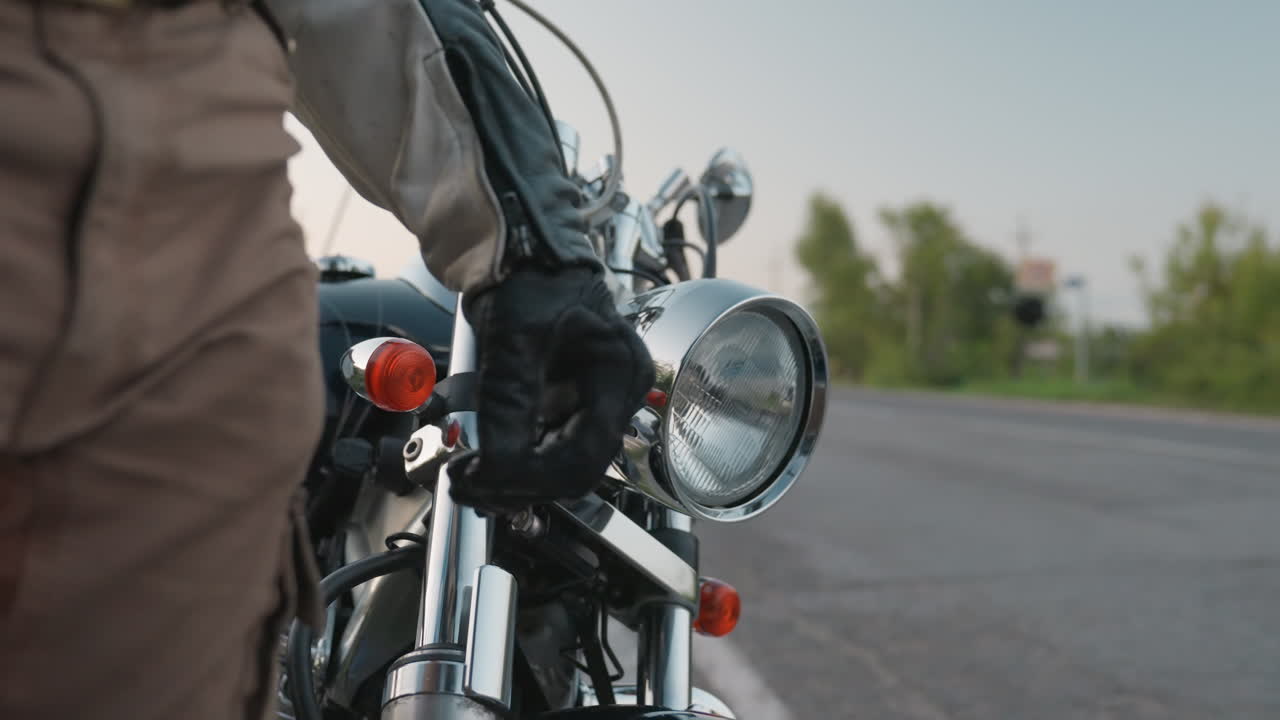 Close up detail of motorcycle handlebar with rider in leather jacket standing beside road during evening ride, highlighting chrome parts, textured gear and lifestyle of freedom