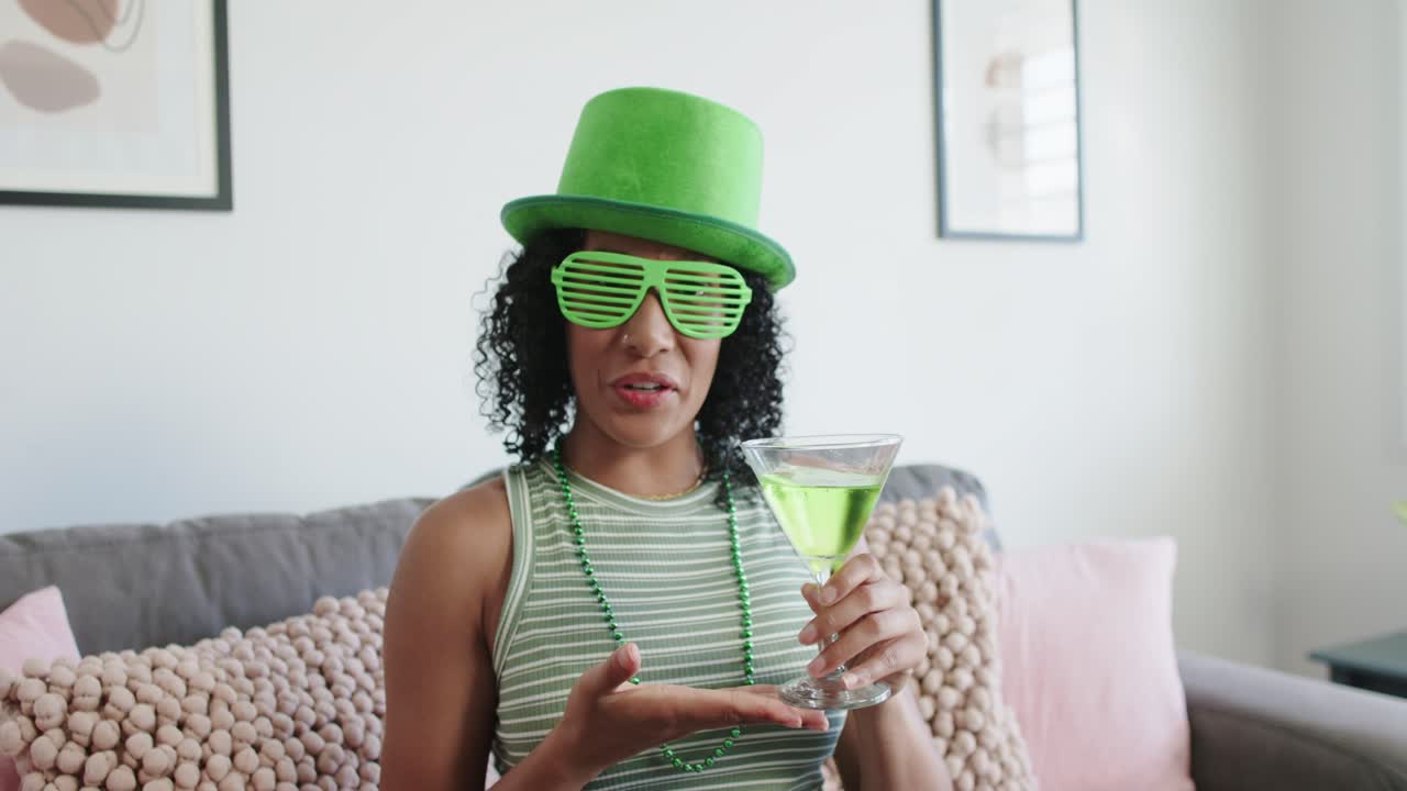 Afroamerikanische Frau berührt lächelnd die Linse, trägt einen grünen Hut und präsentiert einen grünen Martini auf dem Sofa.