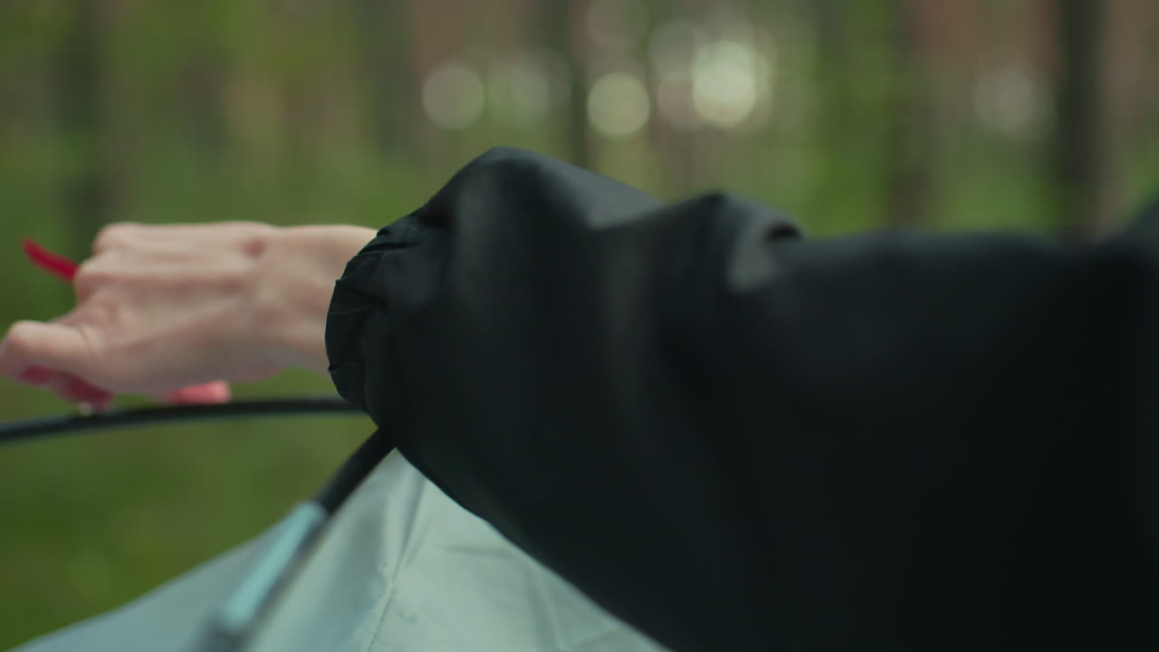close up of lady hand tying red rope of camping tent to flexible black pole while setting up shelter in green forest environment wearing black jacket with soft daylight in background