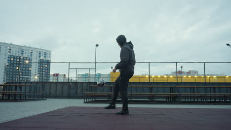 man skillfully juggling football on sport arena with urban background featuring benches, lit pathways, and tall residential buildings