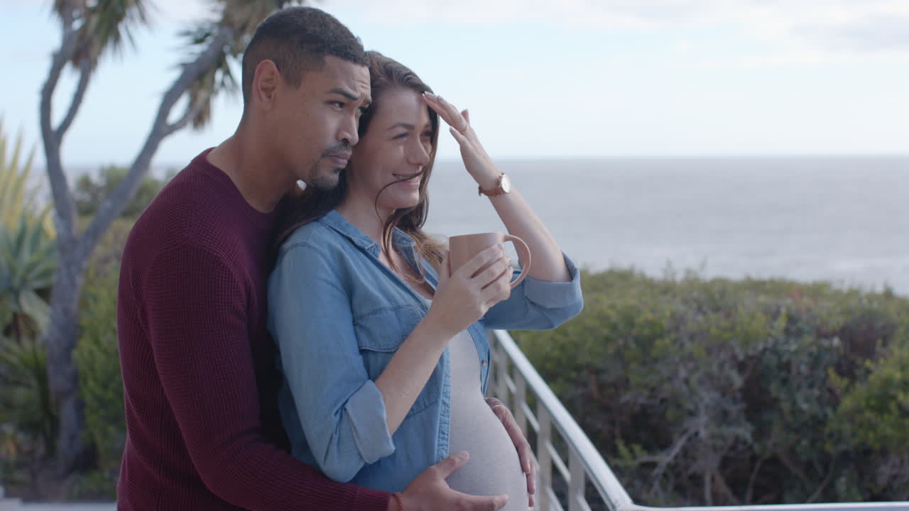 Smiling pregnant couple embracing on balcony, woman holding coffee mug