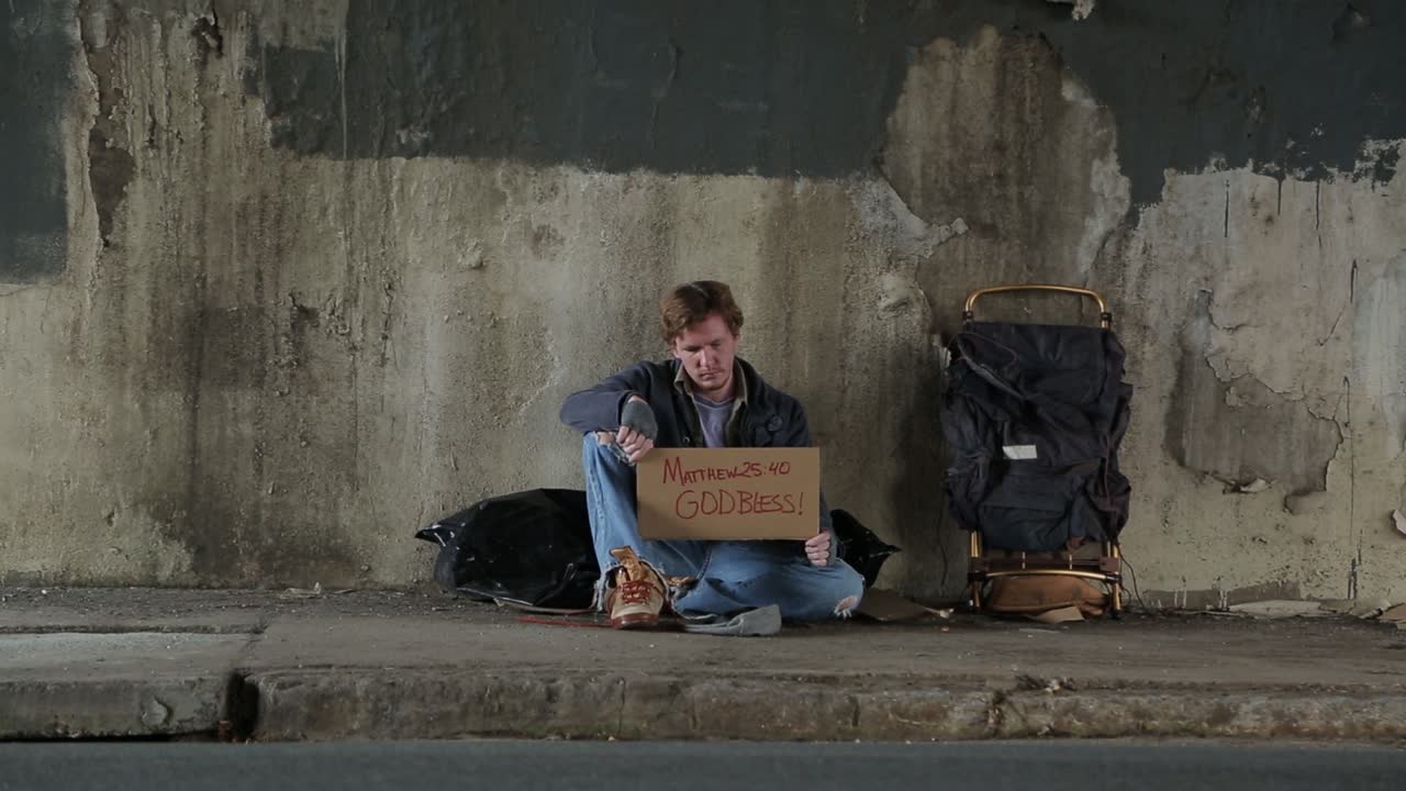 성경 구절과 신의 축복이 있는 표지판을 들고 있는 보도 아래 보도에 있는 노숙자의 와이드 샷