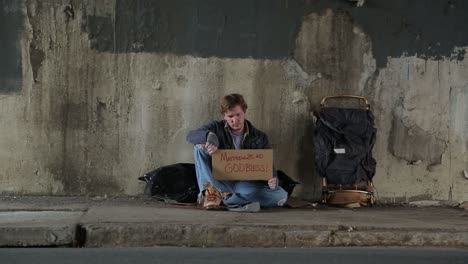 plano general de un vagabundo en la acera bajo un paso elevado sosteniendo un cartel con un verso de la biblia y que dios los bendiga