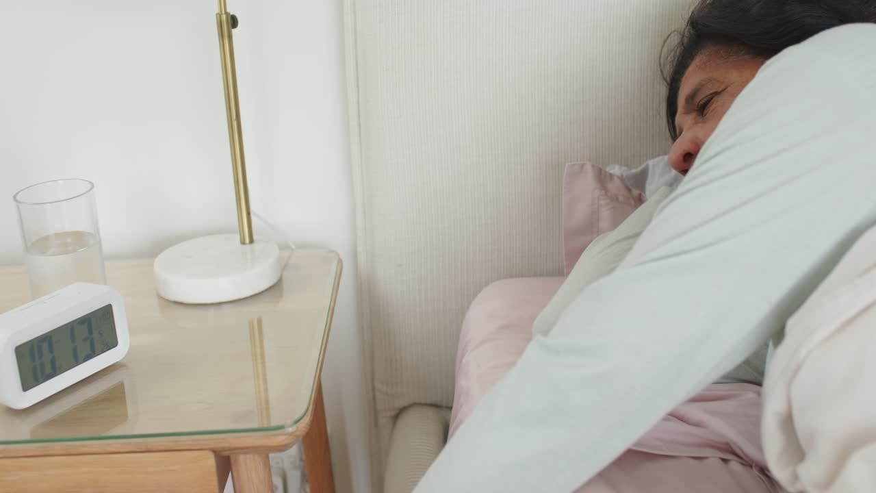 Donna afroamericana matura che tocca la sveglia per farla smettere di suonare, poi riposa a letto, con spazio per il testo.