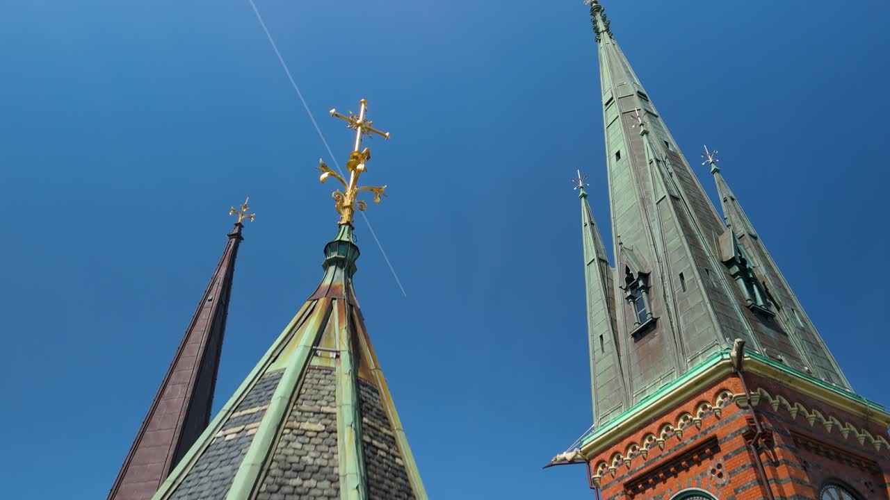 Drone si alza oltre le guglie ornate, la croce dorata e la torre dell'orologio della chiesa gotica in mattoni rossi di Oscar Fredriks Kyrka contro un cielo azzurro vivido a Göteborg
