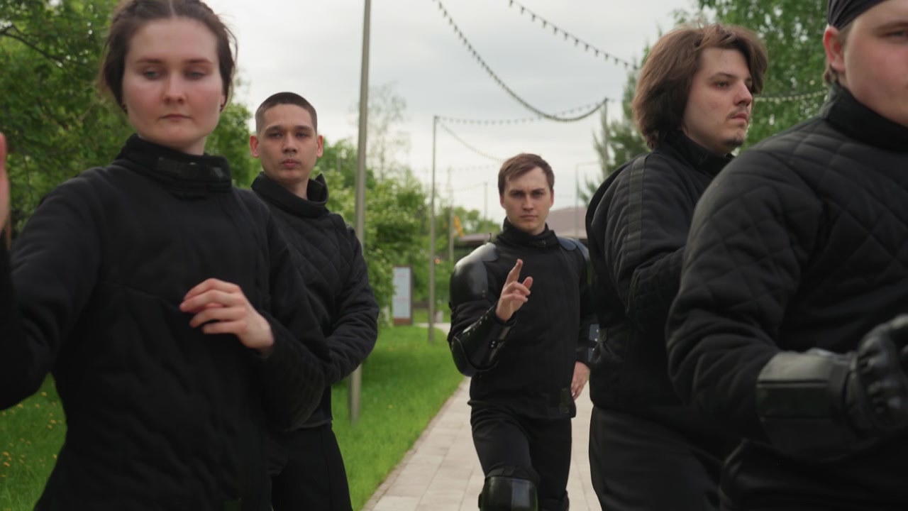 Close up of man in black protective gear walking forward on pavement surrounded by others. man has arm and knee guards visible as they move through outdoor setting with greenery and overcast