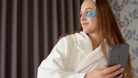 holding smartphone, woman in bathrobe with eye patches relaxing at home spa, copy space