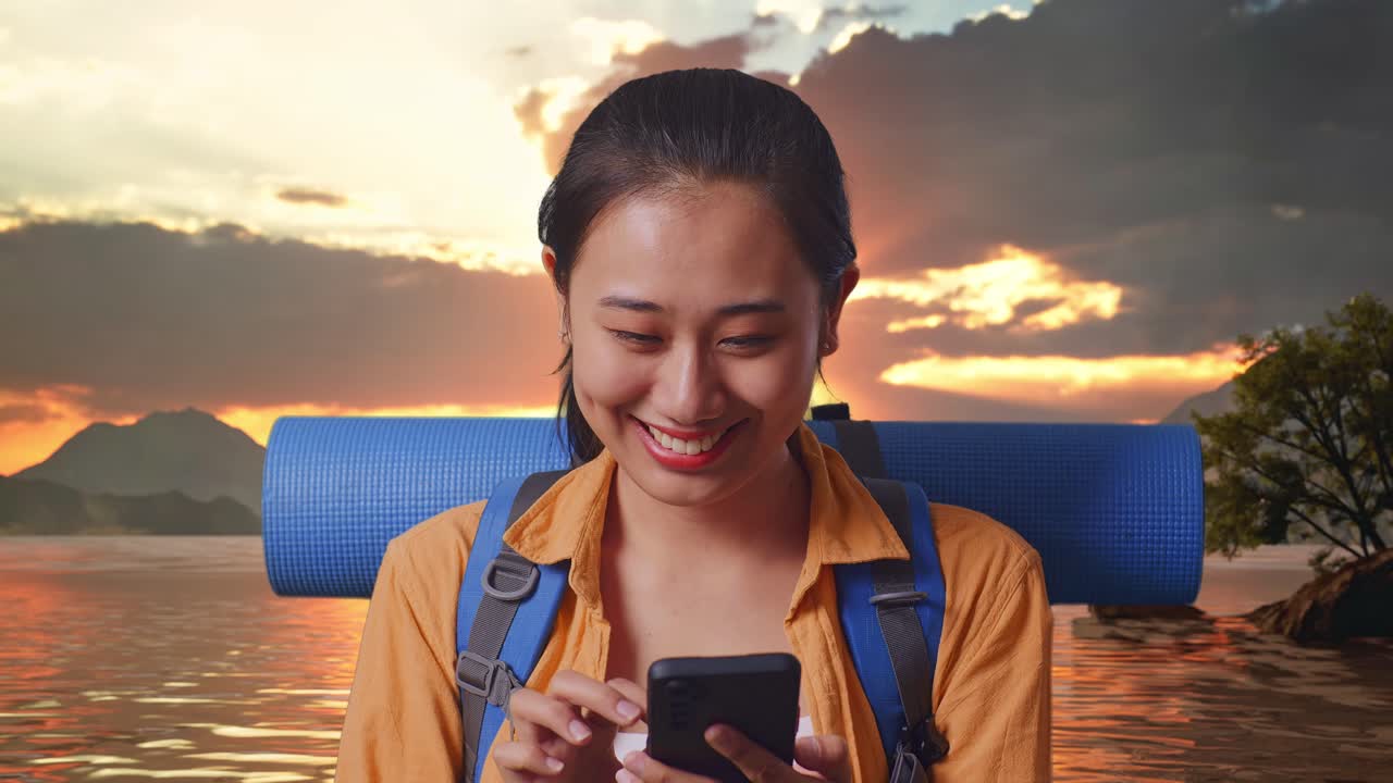 un primer plano de una excursionista asiática con una mochila de montañismo disfrutando de usar un teléfono inteligente mientras está de pie en un lago durante la puesta de sol