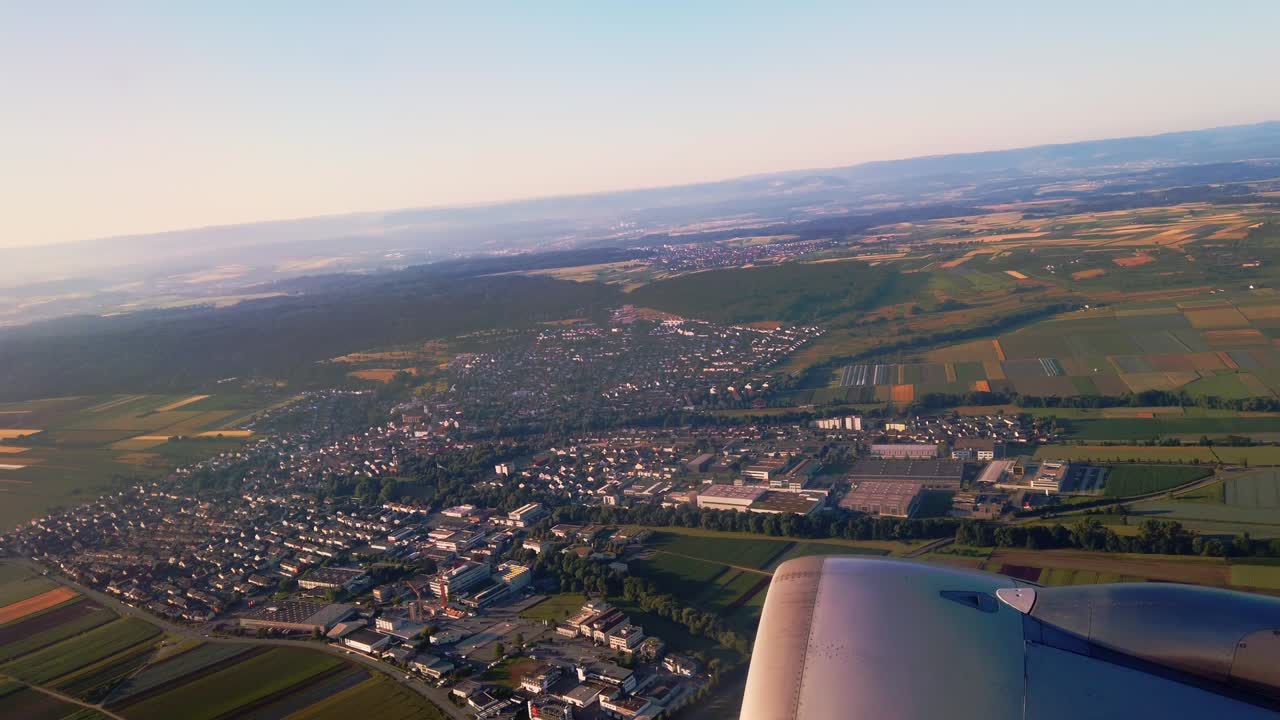 A wide aerial shot from a commercial airplane captures a patchwork of European farmlands, small towns, and distant hills under a clear sky.