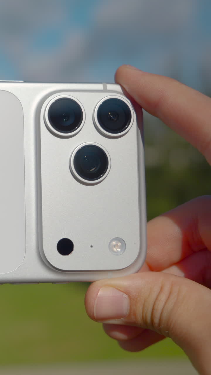 Mani maschili che tengono un prototipo di un nuovo smartphone argento con tre fotocamere, su uno sfondo di parco.
