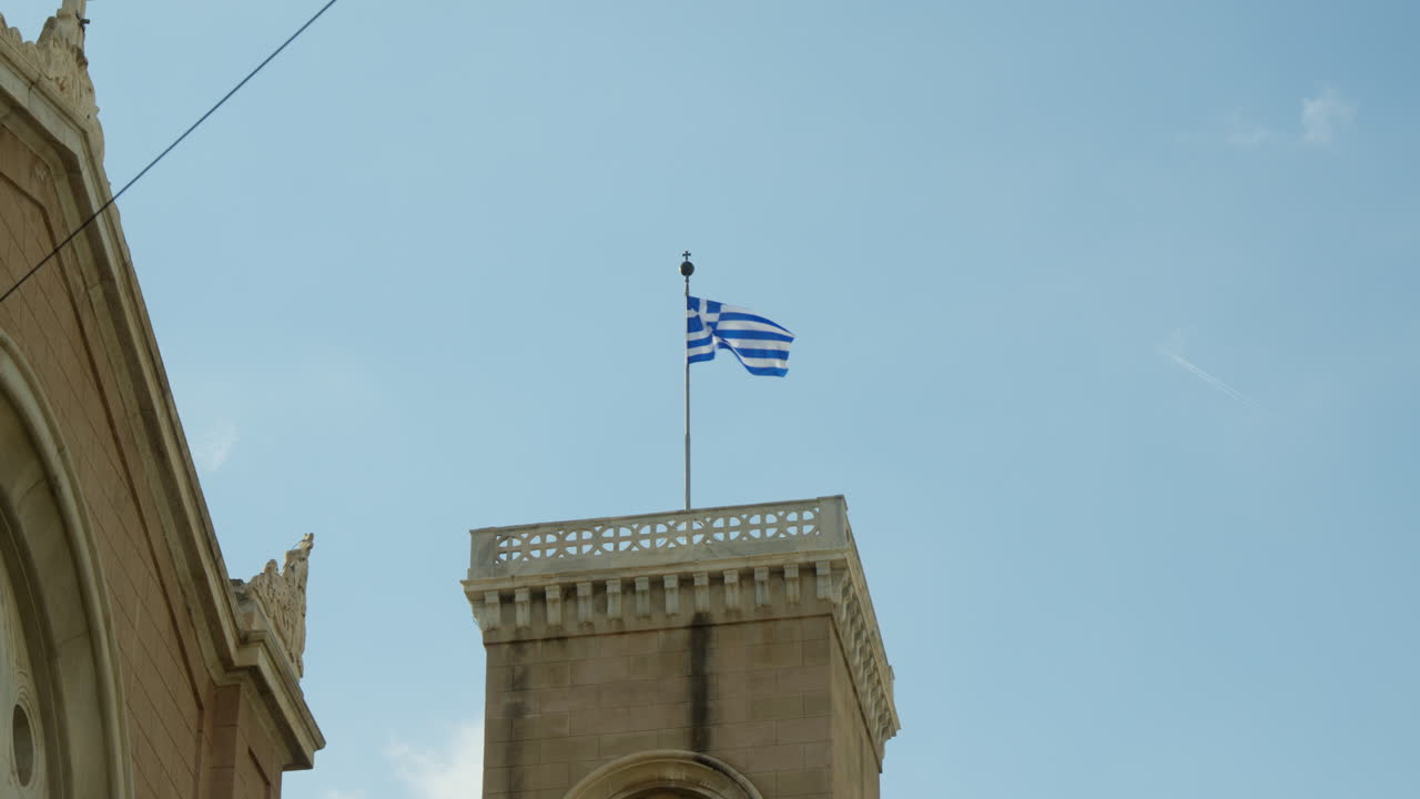 Greek flag waving on historic building's tower against blue sky