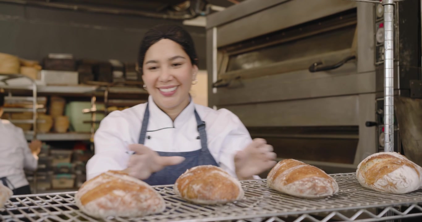 Panettiera di mezza età in giacca in panetteria dopo aver raffreddato le pagnotte, che seleziona il pane e prepara l'impasto
