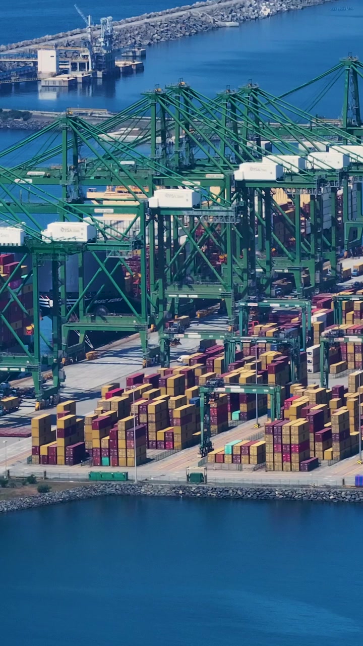 A container terminal where numerous shipping containers are organized. Container ships are being loaded and unloaded as freight moves in and out.