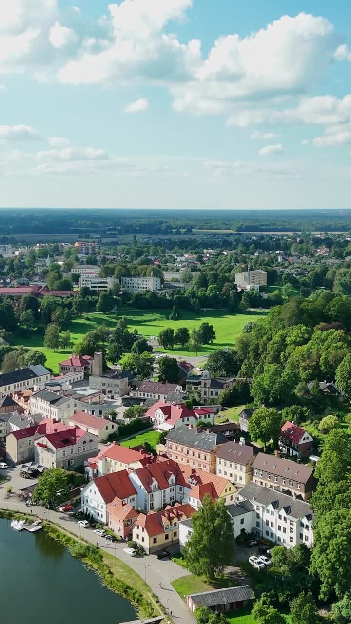 Vertical Aerial panorama capturing Talsi's historic center, featuring red tiled roofs, tranquil waterfront, verdant surroundings, and meandering path under summer sunlight