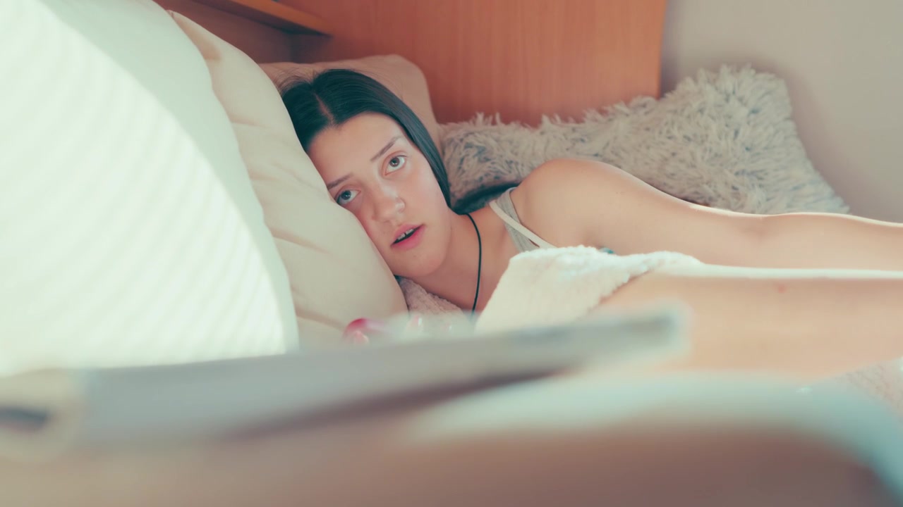 週末の暖かい日差しが差し込むリビングで、ふかふかのソファに横たわる若い女性が、街の友人との交流よりもスマートフォンに夢中になっている様子が捉えられています