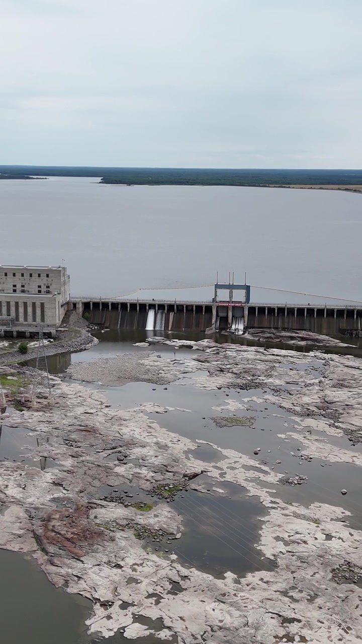 Volo verticale ad alta quota di drone che traccia a destra di fronte alla centrale elettrica Seven Sisters, mostrando rocce esposte dello Scudo Canadese in un canale di sfioro con poca acqua, lago artificiale dietro la diga.