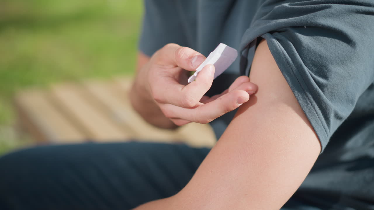 close up of man sitting on wooden bench lifting sleeve to apply nicotine patch on upper arm under bright daylight with soft blur of outdoor greenery in background