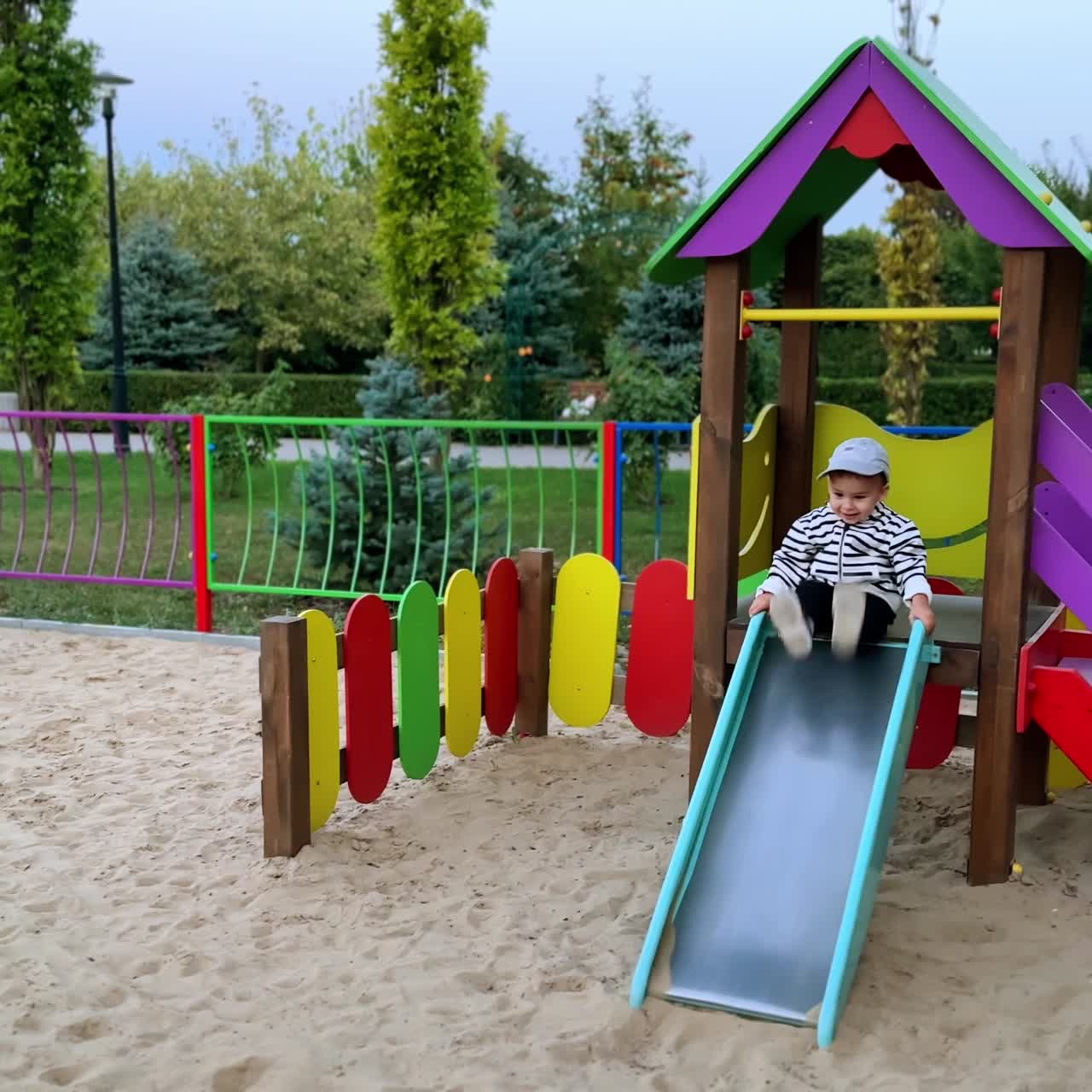 Bebé sonriente con chaqueta bomber a rayas en la diapositiva. Niño moviendo los pies cerca de la diapositiva y luego bajando.