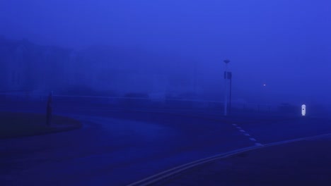 very foggy morning in uk. dark blue light. street lights are still on. car with headlights still on is driving down an empty road and turns towards the camera, drives past. footage in hd