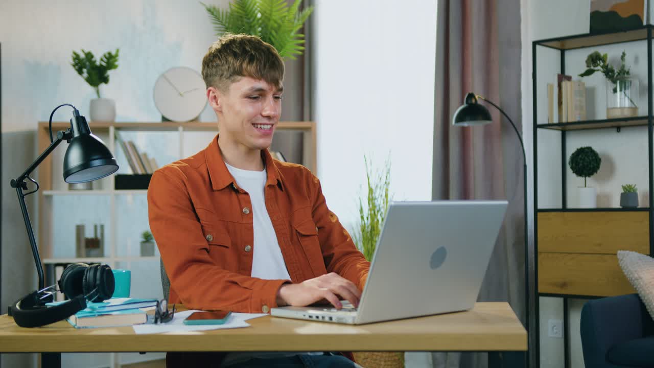 joven rubio guapo sonriente despreocupado con ropa de moda sentado frente a la computadora portátil durante el chat de video con un amigo en casa, concepto de ocio