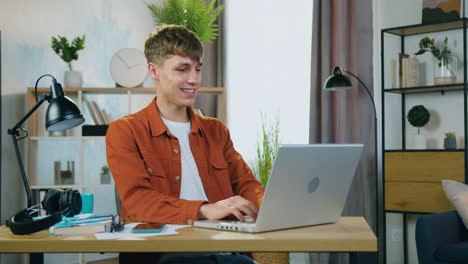 joven rubio guapo sonriente despreocupado con ropa de moda sentado frente a la computadora portátil durante el chat de video con un amigo en casa, concepto de ocio