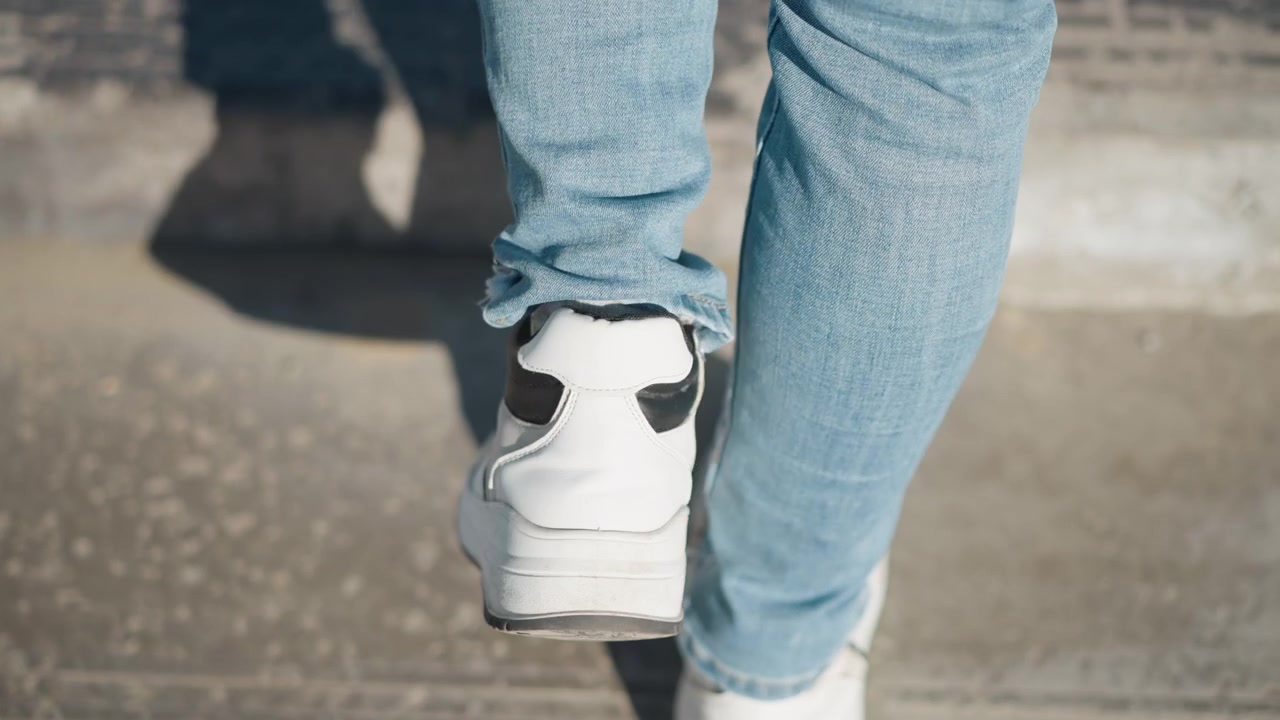 Donna cammina lungo un sentiero di cemento indossando jeans azzurri chiari e scarpe da ginnastica bianche. L'attenzione rimane sulle sue gambe inferiori mentre la donna avanza costantemente in un ambiente esterno con la luce naturale del giorno che proietta