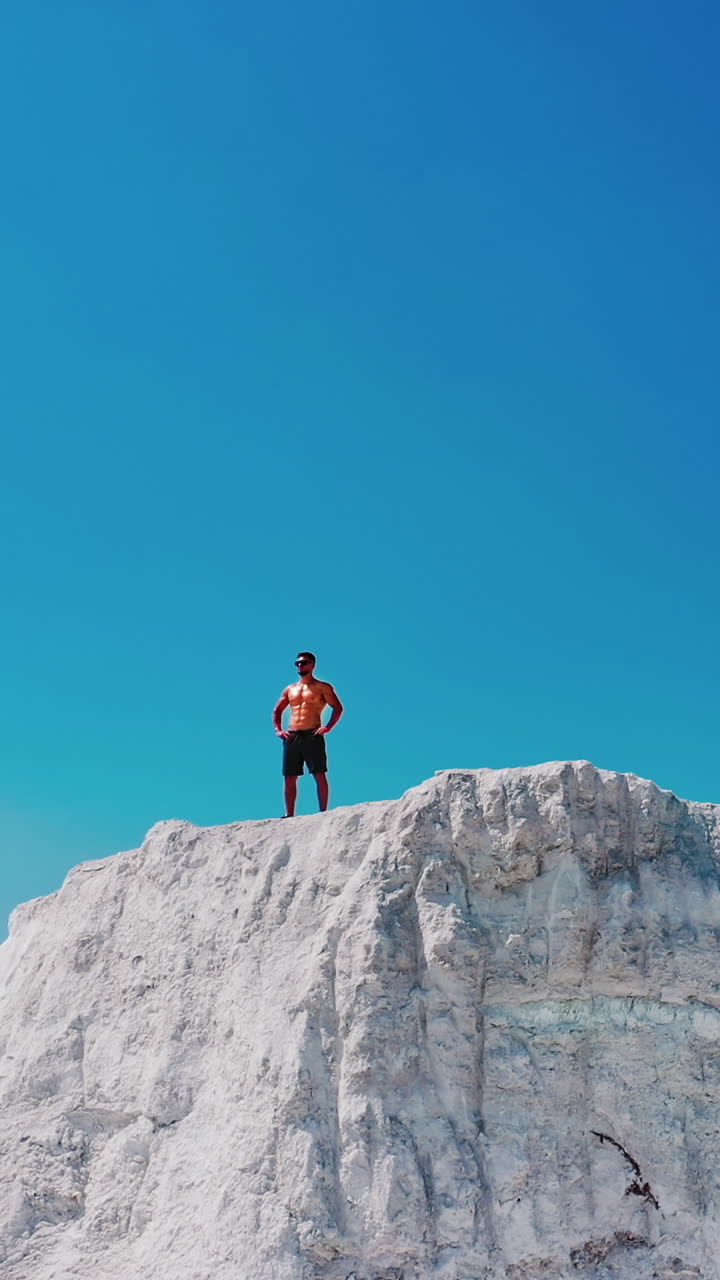 Drone view on a sportive man standing on the edge of the mountain. Shirtless athlete on the top of white canyon under clear sky in summer day. Vertical video