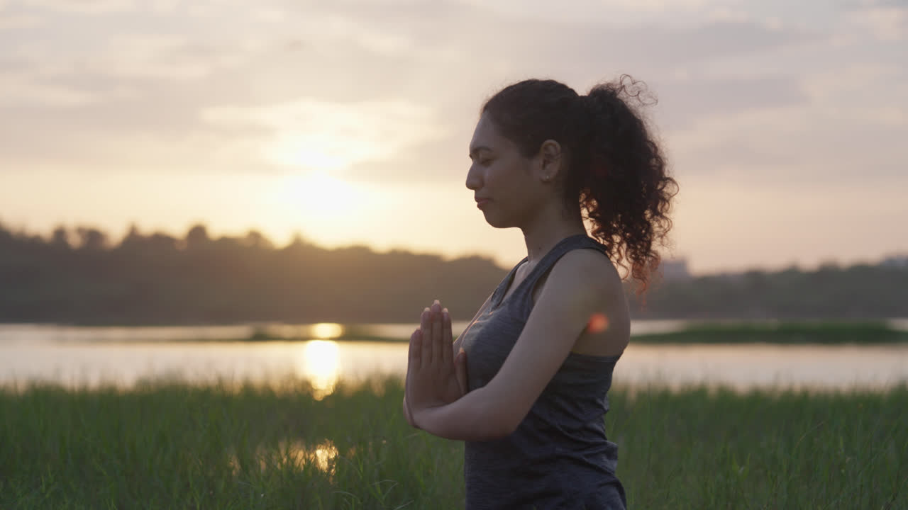 Primo piano di una donna dell'Asia meridionale in posa di preghiera (Anjali Mudra) che fa yoga vicino a un lago al tramonto nell'erba alta