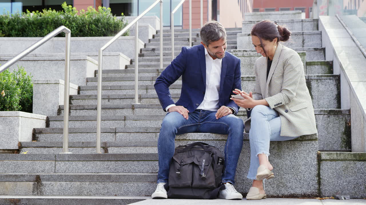hombre de negocios y mujer de negocios sentados por los escalones teniendo una reunión al aire libre mirando el teléfono