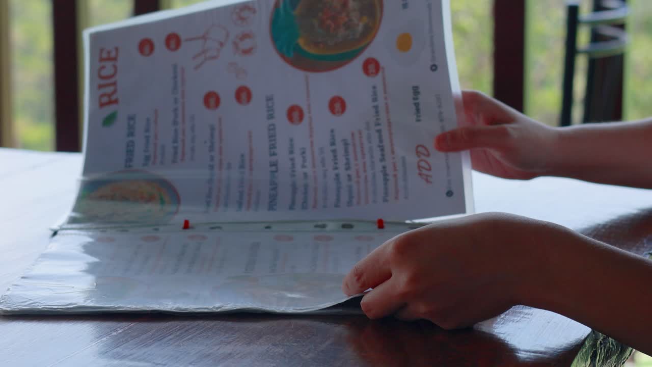 Hands flipping through a menu at an outdoor restaurant in Phuket, Thailand. Natural lighting and a relaxed atmosphere