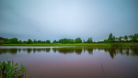 Timelapse-of-Low-Angle-View-of-Tranquil-Pond-Surrounded-by-Lush-Green-Fields-and-Overcast-Clouds-Strolling-By,-with-Changing-Light-and-Vibrant-Colours