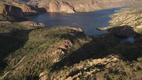 Drone-flying-over-mountains-and-then-slowly-panning-up-revealing-a-dark-blue-lake-with-boats-on-it,-late-evening,-golden-hour,-apache-lake