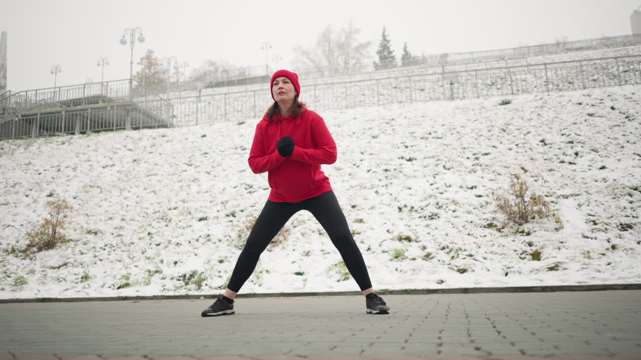 entrenador con capucha roja realiza ejercicio de lanzamiento lateral durante el entrenamiento de invierno al aire libre en un terreno cubierto de nieve cerca de la zona urbana, mostrando determinación, el fondo incluye colina nevada y poste de luz