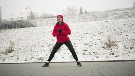 coach in red hoodie performs side lunge exercise during outdoor winter workout on snow-covered ground near urban area, displaying determination, background includes snowy hill and light pole