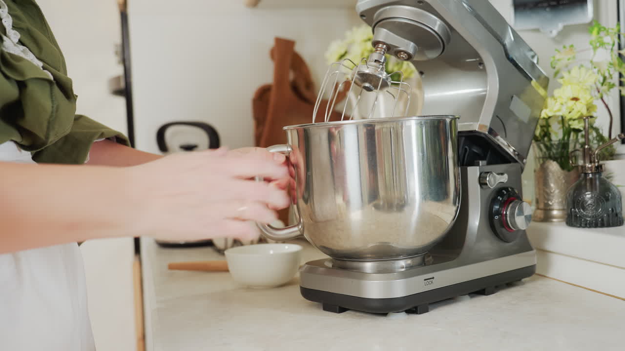 Cook hand placing stainless steel mixing bowl back into electric stand mixer inside cozy kitchen with countertop appliances, white ceramic bowl, and floral decor for food preparation process