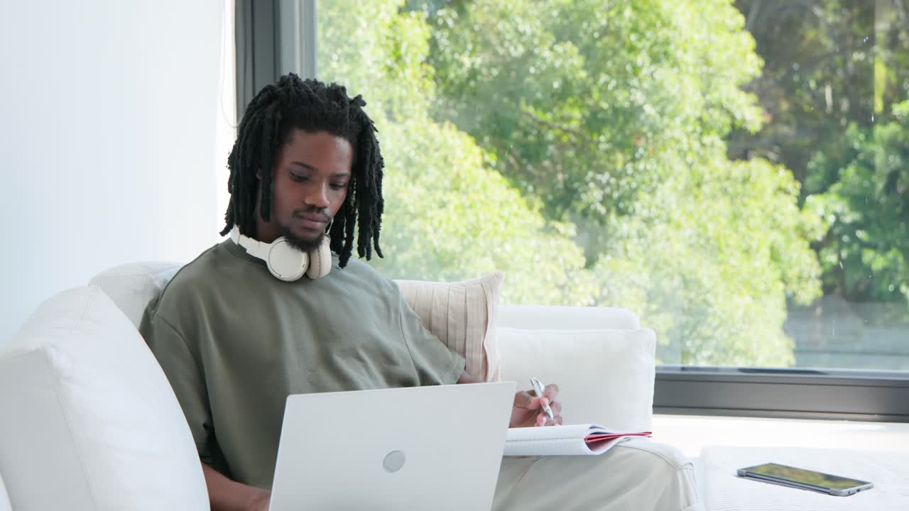 African American man prompted by laptop, checking and writing in red notebook with pen near window
