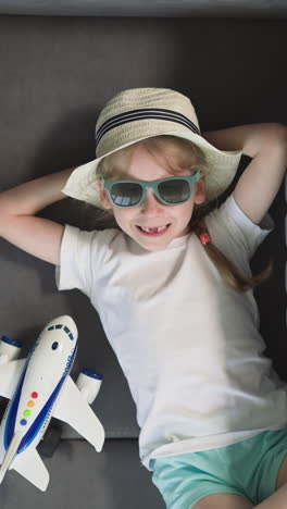 smiling toothless little girl in sunglasses rests on sofa near airplane and suitcase at home. happy preschooler ready for summer vacation upper closeup