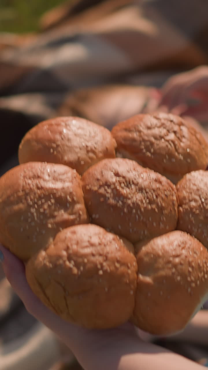 A woman s hand in a blue dress holds a cluster of freshly baked sesame seed bread rolls, gently placing them on a blanket in a grassy field. Her children are visible,slightly blurred in the background