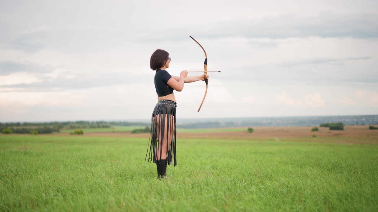 una donna bianca arciere tende l'arco mirando attraverso un campo verde aperto, paramano in pelle visibile, gonna con frange che si muove con il movimento, sequenza dalla trazione al rilascio, respiro costante e postura concentrata, allenamento